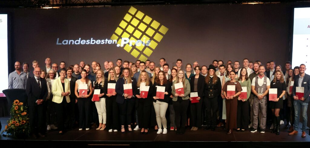 Gruppenfoto auf einer Bühne bei einer Preisverleihung: Etwa 70 Personen stehen nebeneinander, viele halten Urkunden in den Händen. Im Hintergrund ist ein gelbes Rasterdesign mit dem Schriftzug ‚Landesbesten-Preis‘ zu sehen. Links steht ein Rednerpult mit Blumenarrangement. Die Personen tragen formelle Kleidung und lächeln in die Kamera.
